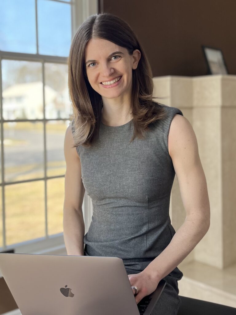 Woman with medium-length light-brown hair wearing a charcoal grey dress with a space grey Macbook pro laptop in her lap. She is smiling and ready to work with a nutrition client.
