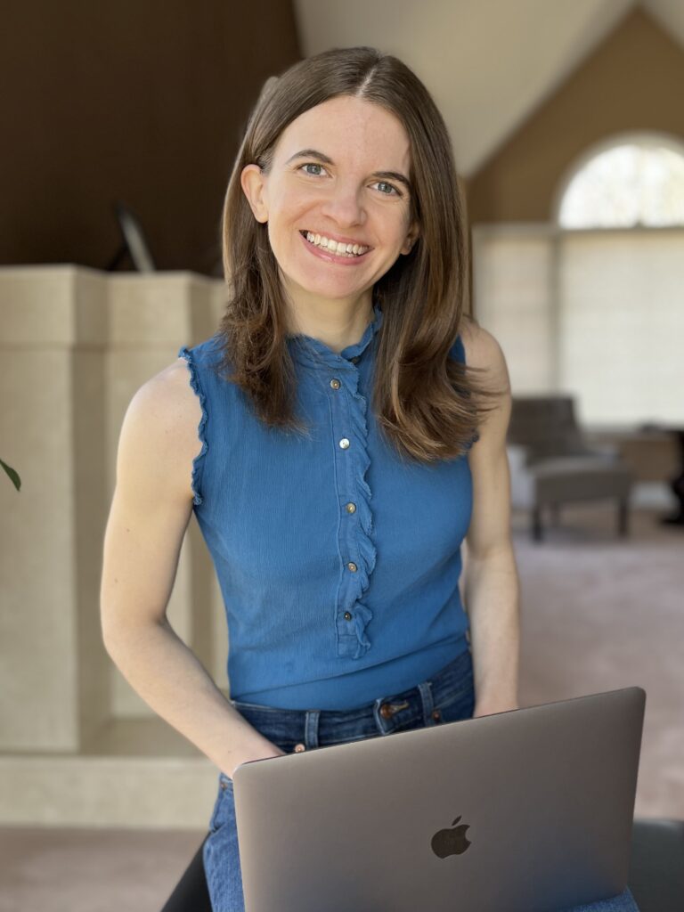 Woman with medium-length light-brown hair wearing a peacock blue sleeveless shirt and blue jeans with a space grey Macbook pro laptop in her lap. She is smiling and ready to work with a nutrition client.