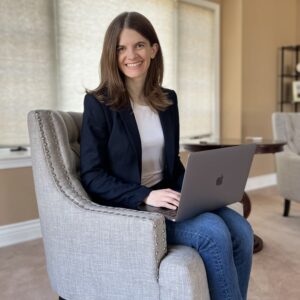 Woman wearing a navy blue blazer, white shirt, and blue jeans sitting in chair with a laptop on her lap. She is smiling at the camera and is ready to work as an online business manager for dietitians.